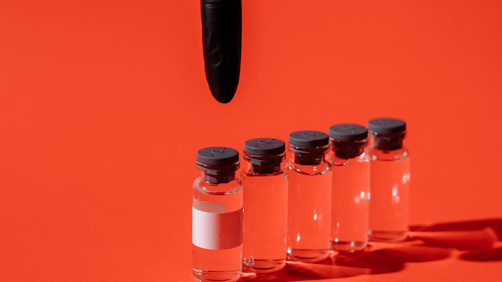 A gloved hand pointing at a row of transparent glass vials arranged against a red background.